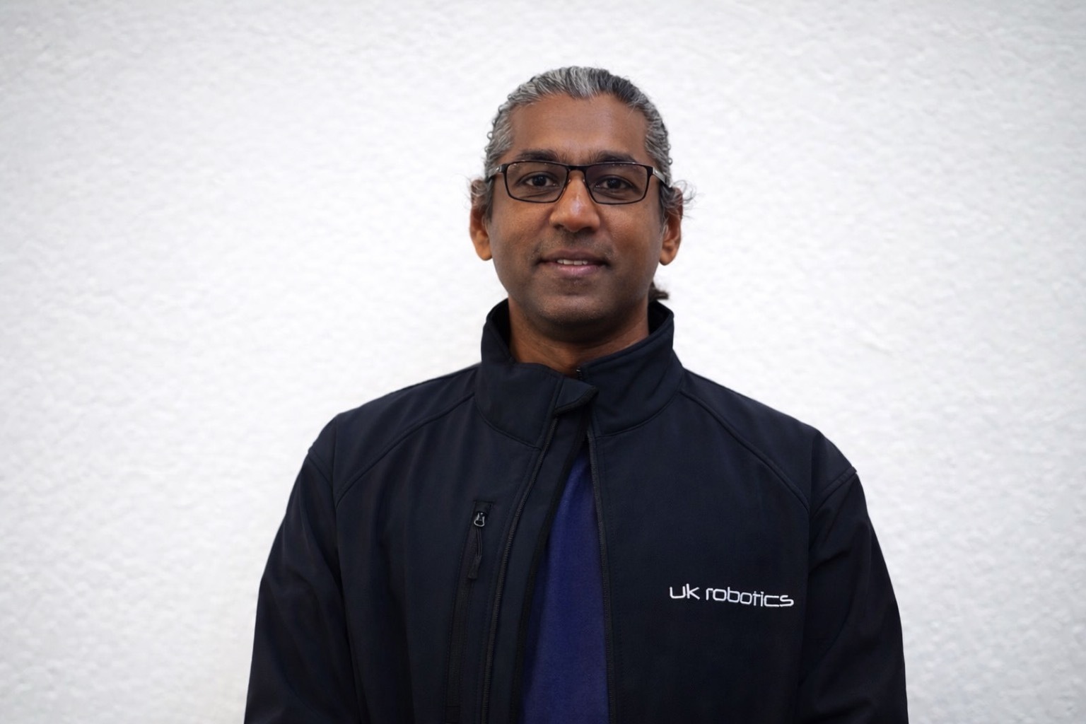 Person wearing a dark jacket with "uk robotics" logo, glasses, against a white wall