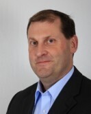 Headshot of a person in a dark blazer and collared shirt against a light background