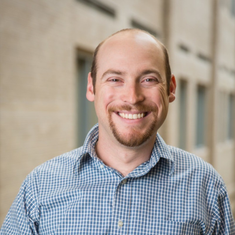 Smiling person with a beard and receding hairline wearing a blue plaid shirt, standing outdoors near a building.